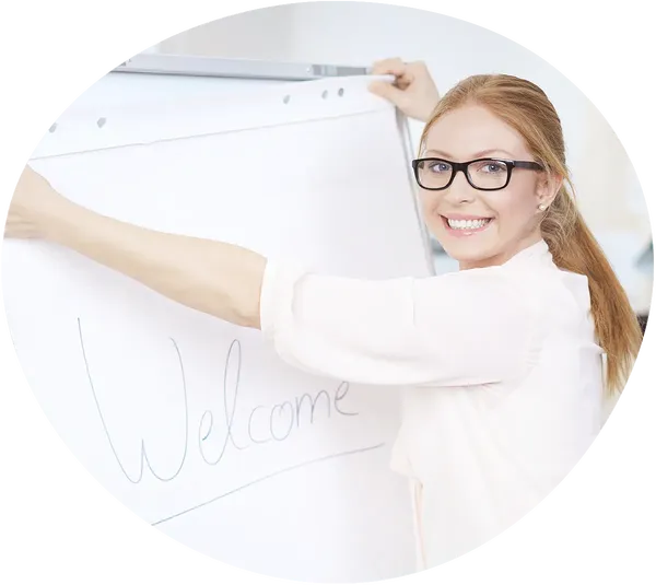 Eine lachende, rothaarige Frau mit Zopf, weißer Bluse und drunkler Brille dreht den Kopf über die Schulter zur Kamera und hängt gerade ein Papierschild mit "Welcome" auf.