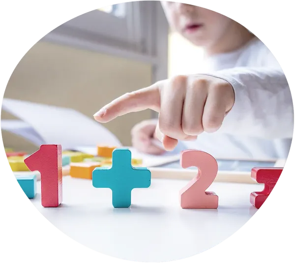 Blick auf einen weißen Tisch auf dem die Zahlen "1 + 2 =" als bunte Holzformen zu sehen sind. Im Hintergrund sitzt ein Kind mit aufgeschlagenem Heft und zeigt auf die 1.