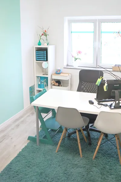 Leicht schräger Blick von oben auf einen Besprechungstisch mit Computer, zwei Stühlen und einem Hochstuhl für ein Kind. Die Sitzgruppe steht vor einem hellen Fenster mit zwei Regalen. Die Wand links ist türlkis.