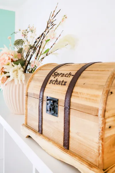 Schatzkiste aus hellem Holz mit Aufschrift "Sprachschatz" steht vor Vase mit beigen und lachsfarbenen Blumen auf einer weißen Ablage.