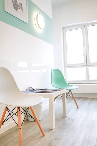 Blick auf zwei Stühle und einen kleinen Tisch m it Zeitschriften auf der linken Seite des Bildes. Im Hintergrund sieht man ein großes Fenster.